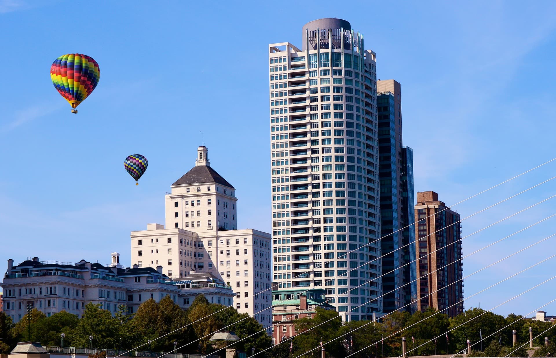 Rainbow and checkered hot air balloons float in a blue sky above a city skyline.